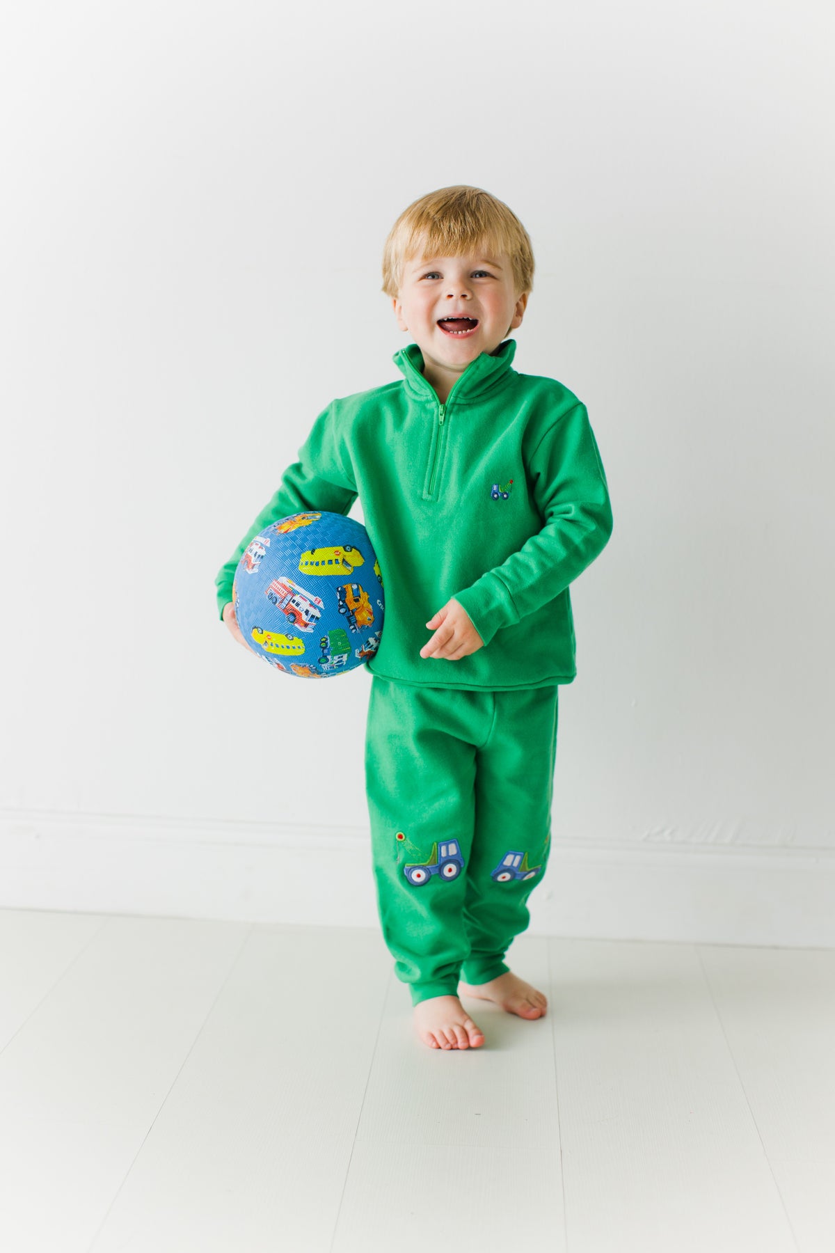 Laughing toddler holding a ball, wearing the green Tow Truck Scout Quarter Zip and Adventure Joggers