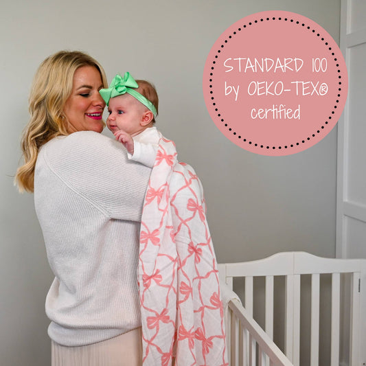 White muslin swaddle blanket with pink bow print, draped over a baby held by their mother in a nursery