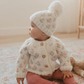 White knit "bitty blooms" pom pom beanie with floral pattern, worn by baby in matching cardigan sitting on a floor rug. 