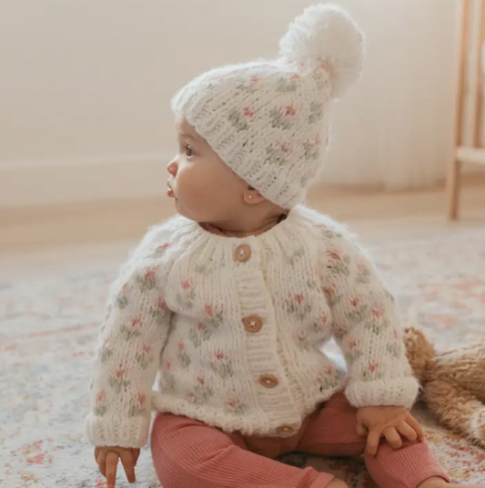 White knit "bitty blooms" pom pom beanie with floral pattern, worn by baby in matching cardigan sitting on a floor rug. 
