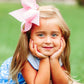 Little girl wearing scallop edged hair bow and blue dress, leans with hands on chin on a box in a garden
