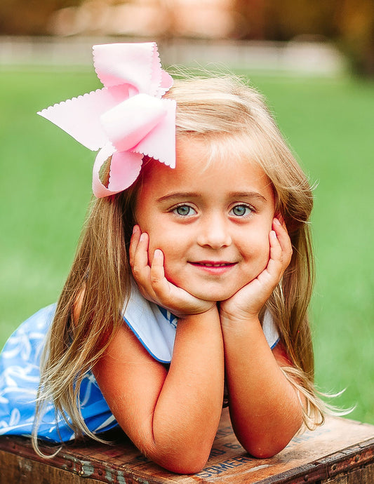 Little girl wearing scallop edged hair bow and blue dress, leans with hands on chin on a box in a garden