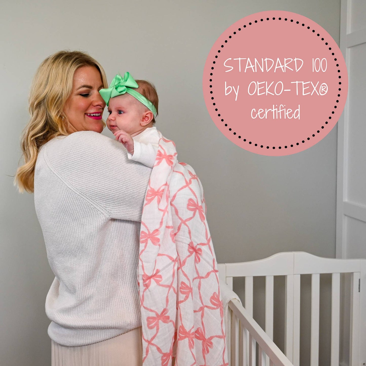 White muslin swaddle blanket with pink bow print, draped over a baby held by their mother in a nursery