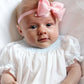 baby girl wearing pink scallop edge headband and white smocked bubble with blue detail, lays on white bed