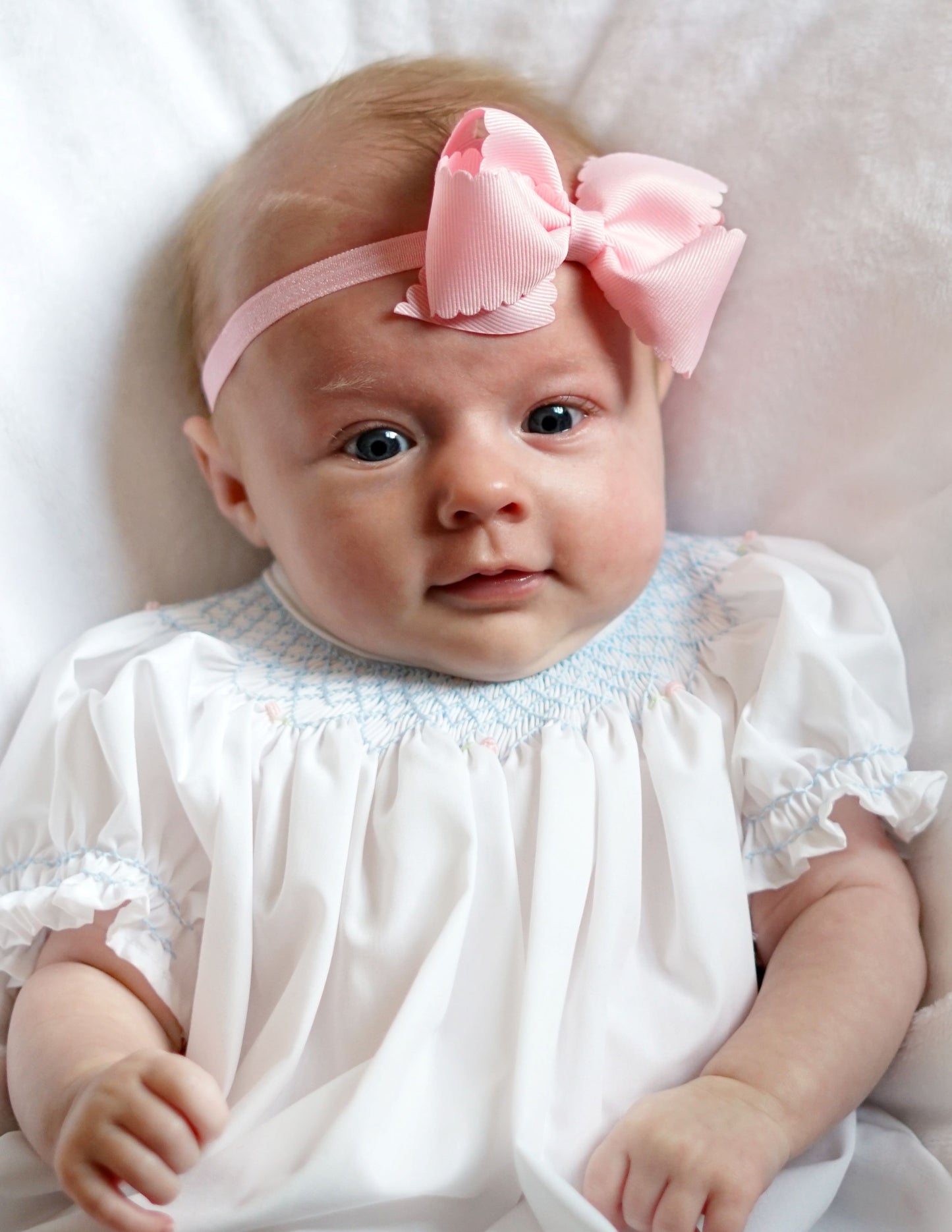 baby girl wearing pink scallop edge headband and white smocked bubble with blue detail, lays on white bed