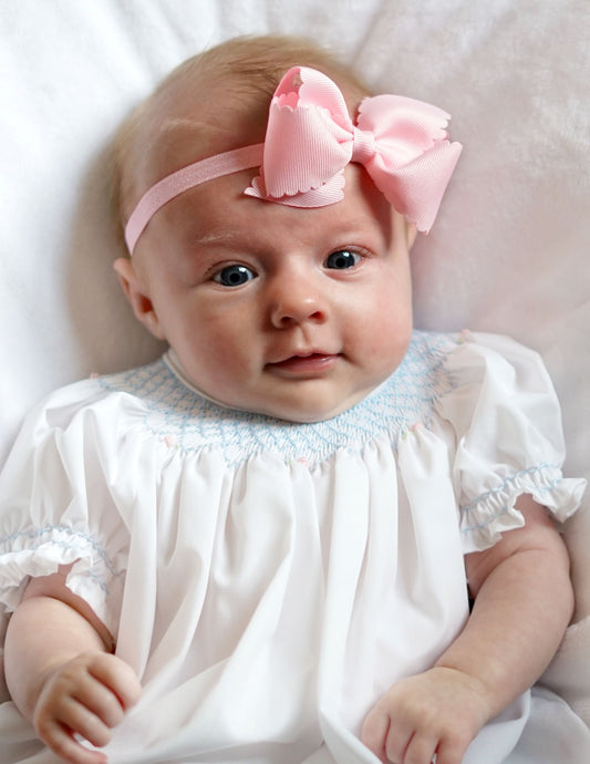 baby girl wearing pink scallop edge headband and white smocked bubble with blue detail, lays on white bed